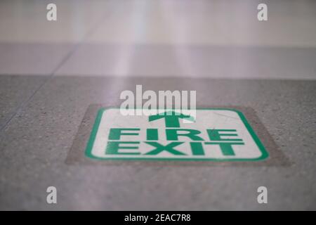A green fire exit with an arrow on a gray tile floor, pointing to a safe zone area outside the building in case of emergency or fire. Stock Photo