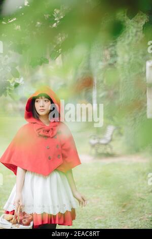 Portrait young woman with Little Red Riding Hood costume with apple and bread on basket in green tree park background Stock Photo