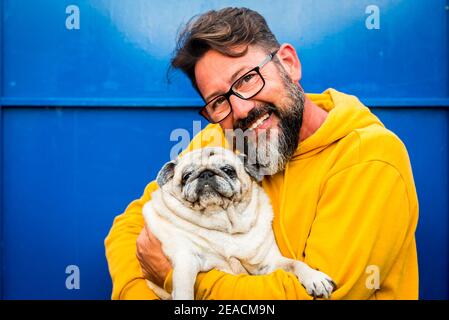 Cheerful adult man smile and hug with love his own old dog pug in a portrait with yellow and blue color - people with animals and glasses enjoy the pet therapy concept Stock Photo