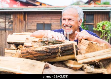 Cheerful senior man work outdoor at home with dry wood ready to fire and warm and heat home in winter season - active retired people and outdor natural lifestyle in country side house Stock Photo