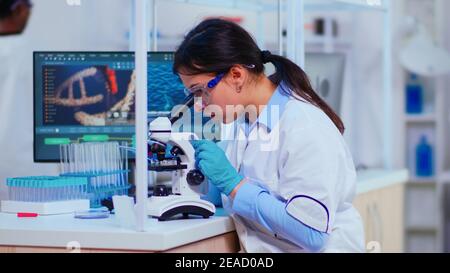 Scientist woman with microscope examining samples and liquid working in modern equipped laboratory. Team of doctors examining vaccine evolution using high tech researching diagnosis against covid19 Stock Photo