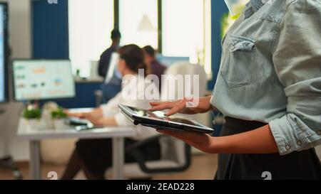Employee using tablet standing in office room while team of financial analysts working in background. Multiethnic coworkers respecting social distance in business company during coronavirus pandemic. Stock Photo