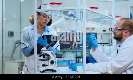Diverse team of biochemistry scientists developing drugs agains new virus, doctor checking samples while nurse taking noters on clipboard in modern equipped laboratory. Multiethnic scientists working Stock Photo