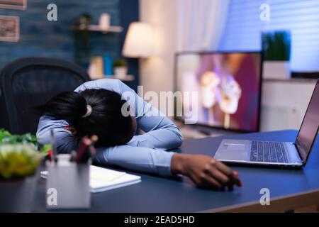 Exhausted dark skinned woman resting on office desk in home office late at night. Busy employee using modern technology network wireless doing overtime sleeping on table. Stock Photo