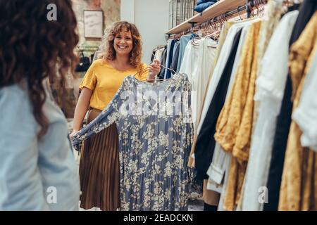 Woman holding a dress and showing it to female customer in her store. Clothing store owner helping a customer in shop. Stock Photo