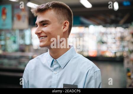 Close-up of a man working in a grocery store. Young worker looking away and smiling in supermarket. Stock Photo