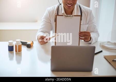 Image of professional meeting on video call while therapist sitting at the table and showing clipboard at the camera of laptop Stock Photo