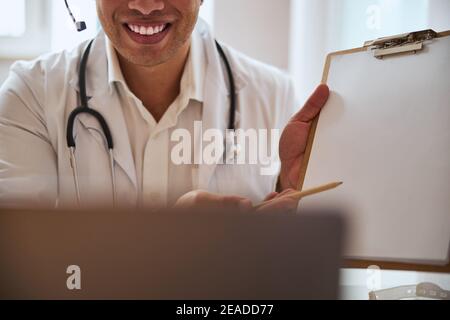 Confident therapist in with stethoscope sitting at the table and having conversation with patient at the epidemic time Stock Photo