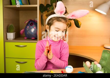 A girl in the ears of an Easter bunny paints eggs with a felt-tip pen in the home interior. Crafts, preparation for a religious holiday, a tray with e Stock Photo