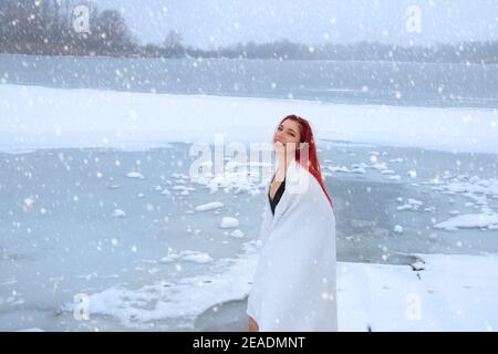 Redhead woman wrapped in a towel with a happy smile enjoying cold therapy on freezing lake, hardening on snow, ice bath, and winter swimming concept