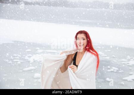 Snowy winter portrait of happy smiling woman in snowfall outside in a bathing suit, wrapped in a towel at open water swimming lake