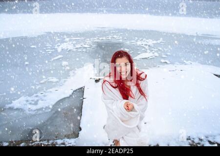 Happy young woman with long red hair, wrapped only in a blanket with a joyful smile enjoys natural cold therapy and snow shower in winter outdoors