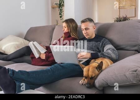 Young couple and their dog enjoying leisure time and relaxing on the couch at home, reading a book and using a laptop. Couple life concept Stock Photo