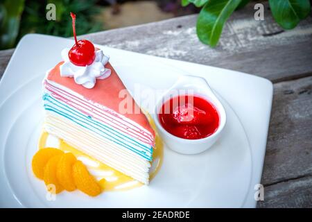 Rainbow crepe cake and a cherry with strawberry sauce on wooden table background. Stock Photo