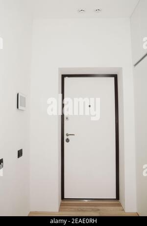 White entrance hall, entryway in a renovated apartment with black and white armored door, a peephole, door handle and door lock, smoke detector. Stock Photo