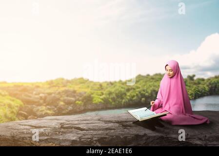 Asian Muslim woman in a veil sitting and reading the Quran at outdoor Stock Photo