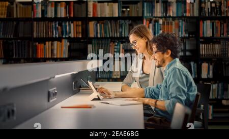 University Library: Cute Couple of Students Have Discussion, use Laptop, Prepare for Exams Together, Helping, Researching Subjects for the Paper Stock Photo