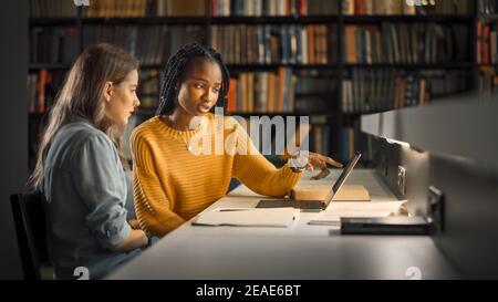 University Library: Two Gifted Girl Students Study, Use Laptop Computer, Help Each Other with Advice, Working on Assignment and Preparing for Exams Stock Photo