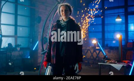 Young Beautiful Empowering Woman with Ear Piercing Gently Smiles at the Camera. Authentic Fabricator Wearing Work Clothes in a Metal Workshop. Sparks Stock Photo