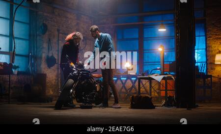 Two Young Professionals are Discussing The Work Done on a Custom Bobber Motorcycle. Hipster Man Explaining the Job to a Talented Female Mechanic Stock Photo