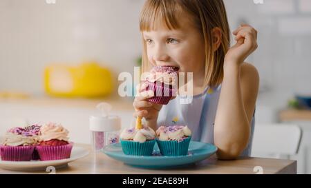 In the Kitchen: Adorable Little Girl Eats Creamy Cupcake with Frosting and Sprinkled Funfetti. Cute Hungry Sweet Tooth Child Bites into Muffin with Stock Photo