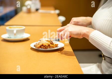 woman cuts croissant and drinks coffee at a table in a cafe. no face Stock Photo