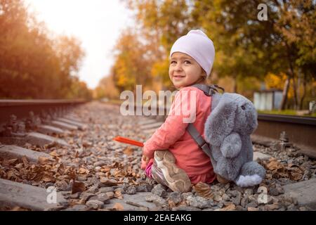 children in danger concept. a cheerful girl sits on the railroad tracks and smiles slyly on sunny autumn day. Stock Photo