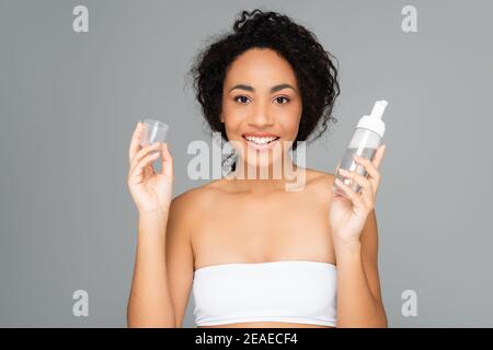Cheerful african american woman holding bottle and cap of cleansing foam isolated on grey Stock Photo