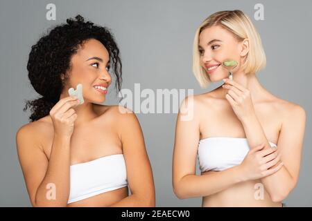 Smiling multiethnic women in white tops using jade roller and gua sha isolated on grey Stock Photo