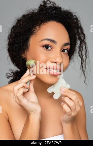 African american woman holding jade roller and gua sha near face isolated on grey Stock Photo