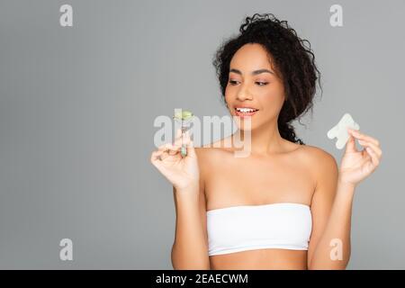 Smiling african american woman in white top holding jade roller and gua sha isolated on grey Stock Photo