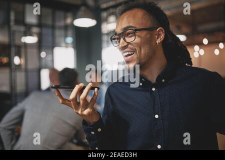Young businessman laughing and talking on speakerphone while standing in an office with colleagues in the background Stock Photo