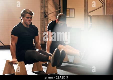 Two fit young men in sportswear doing l-sits during a workout with parallettes on the floor of a gym Stock Photo