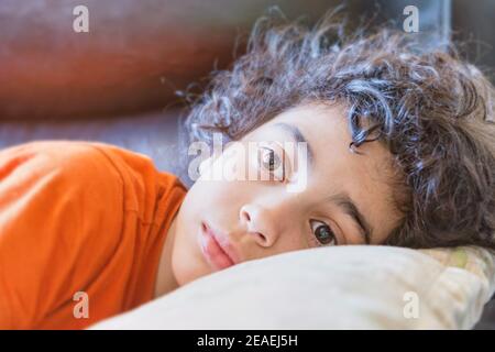 Latin toddler boy with curly hair lying down on a black couch with his white pillow. Stock Photo