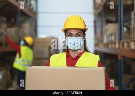 Latin woman working in warehouse carrying delivery boxes while wearing face mask during corona virus pandemic - Logistic and industry concept Stock Photo