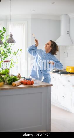 Vertical Shot of Energetic Young Man with Long Hair Dancing in the Kitchen while Wearing Blue Pajamas. Bright White Modern Kitchen Area with Healthy Stock Photo