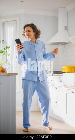 Happy Young Man with Long Hair Dancing at Home while Wearing Blue Pajamas. He is Listening to Music on a Mobile. Energetic Man Using Smartphone in a Stock Photo
