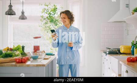 Happy Young Man with Long Hair is Using Smartphone at Home while Wearing Blue Pajamas. He is Holding Cup of Coffee. Energetic Man Scrolling News Feed Stock Photo