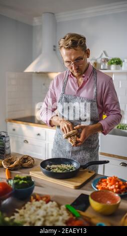 Handsome Young Man Preparing a Healthy Vegetarian Meal on a Frying Pan. Sprinkling Healthy Green Food with Herbs and Spices in a Modern Kitchen Stock Photo