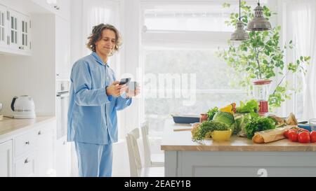 Happy Young Man with Long Hair is Using Smartphone in a Kitchen while Wearing Blue Pajamas. He is Scrolling Social Meadia and News Feed. Energetic Man Stock Photo