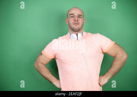 Young bald man using headphones over green background skeptic and nervous, disapproving expression on face with arms in waist Stock Photo