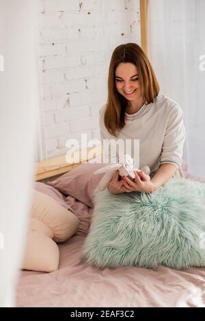 Happy woman holding rabbit toy relaxing on comfortable bed with pillow. Lady with handmade playtoy Stock Photo