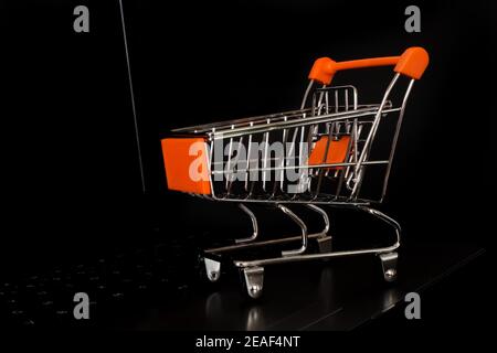 Orange mini shopping cart on the laptop keyboard. Online shopping concept that enables people to shop easily and quickly. Black background. Stock Photo