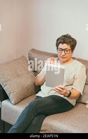 Attractive elegant senior woman sitting on the couch and using a tablet computer at home. Stock Photo