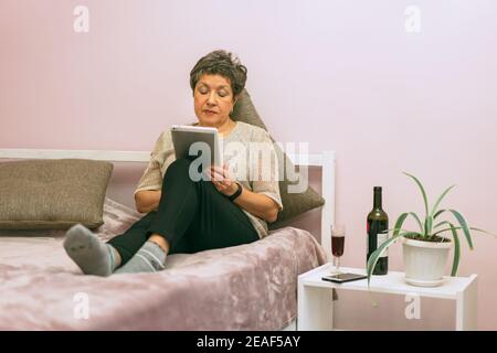 Attractive elegant senior woman sitting on the bed and using a tablet computer at home. Stock Photo