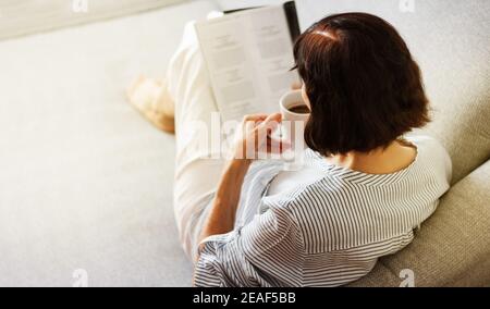 Middle-aged brunette woman with glasses on the gray sofa reading magazine with cup of coffee, soft focus Stock Photo