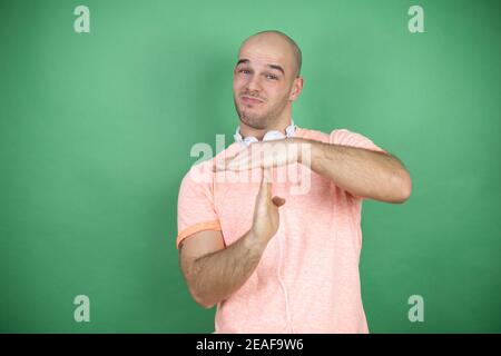 Young bald man using headphones over green background Doing time out gesture with hands, frustrated and serious face Stock Photo