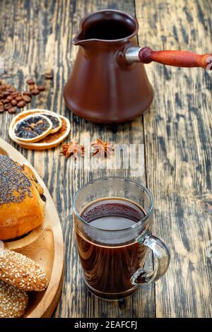 A freshly made espresso coffee in a clear glass cup stands next to a muffin on an old striped wooden table. In the background there is a brown teapot, Stock Photo