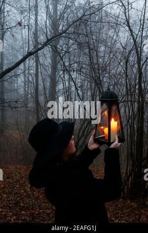 Young woman walks through the dark forest, she is holding an iron lantern. Foggy landscape in the background. Stock Photo
