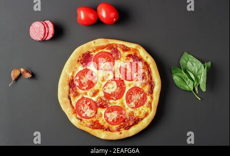 Pizza with sausage, tomatoes and cheese and ingredients around it on a black background. View from above Stock Photo